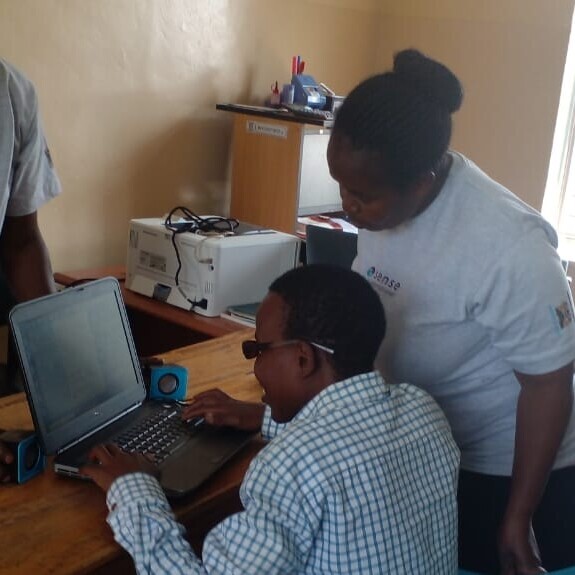 A young person working at a computer with a woman standing nearby